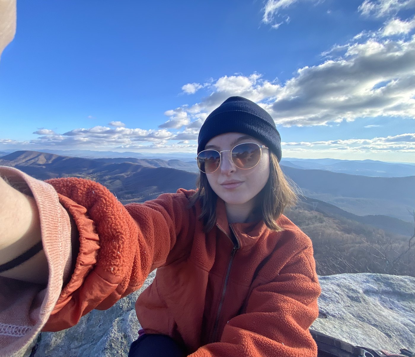 Madeline in a beanie and sunglasses on a summit with mountains behind