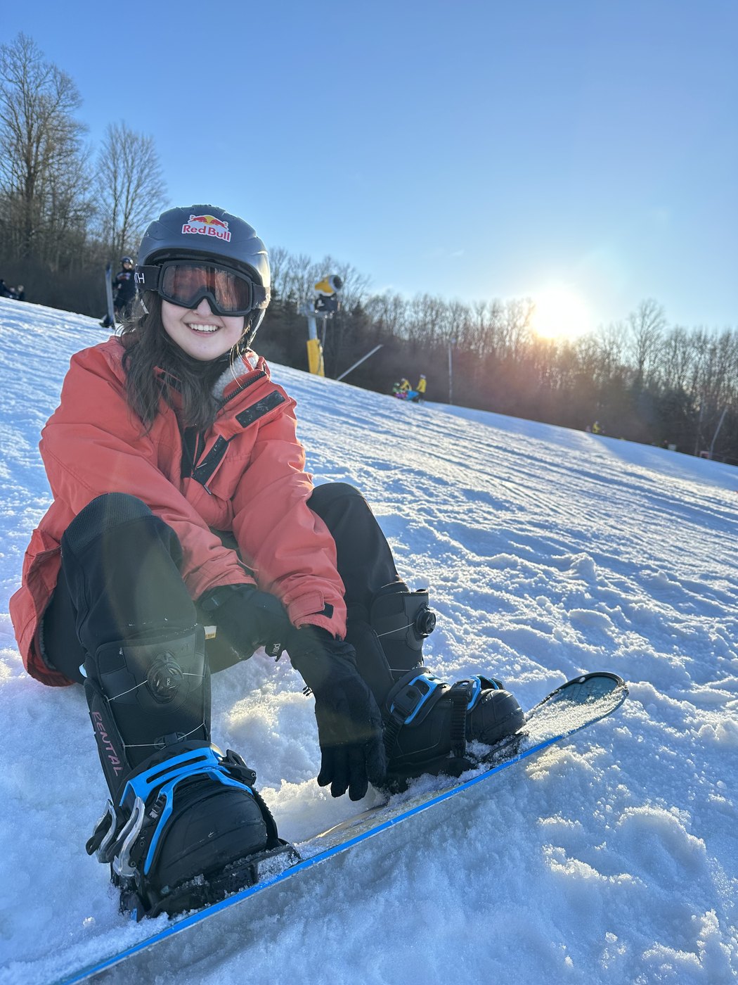 Madeline sitting strapped into a snowboard at the top of a sunlit slope