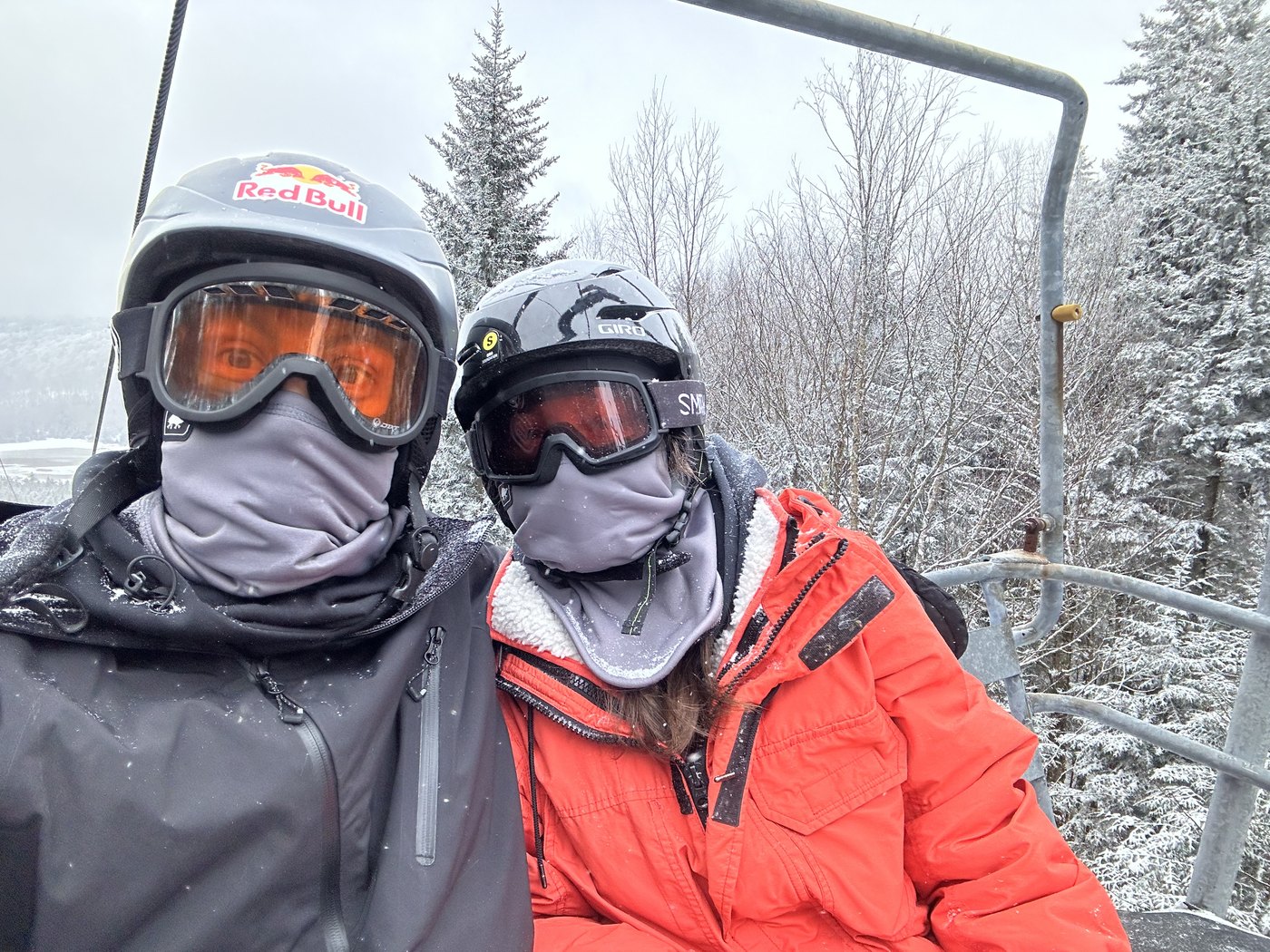 Two snowboarders in goggles and face coverings riding a chairlift through snowy pines