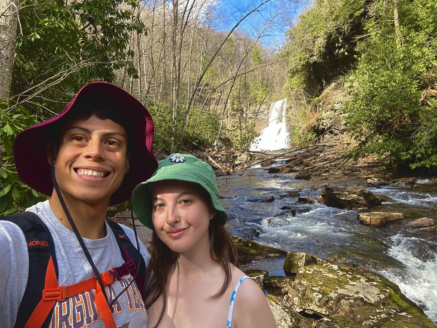 Two hikers smiling beside a rocky creek below a waterfall