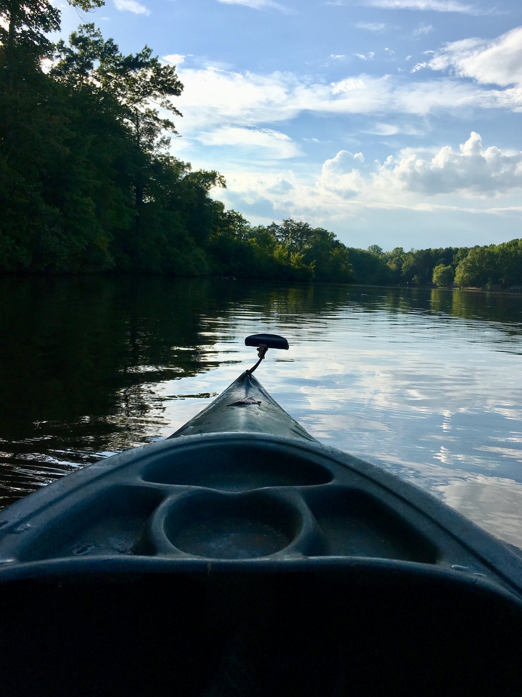 The bow of a kayak on flat water lined with trees