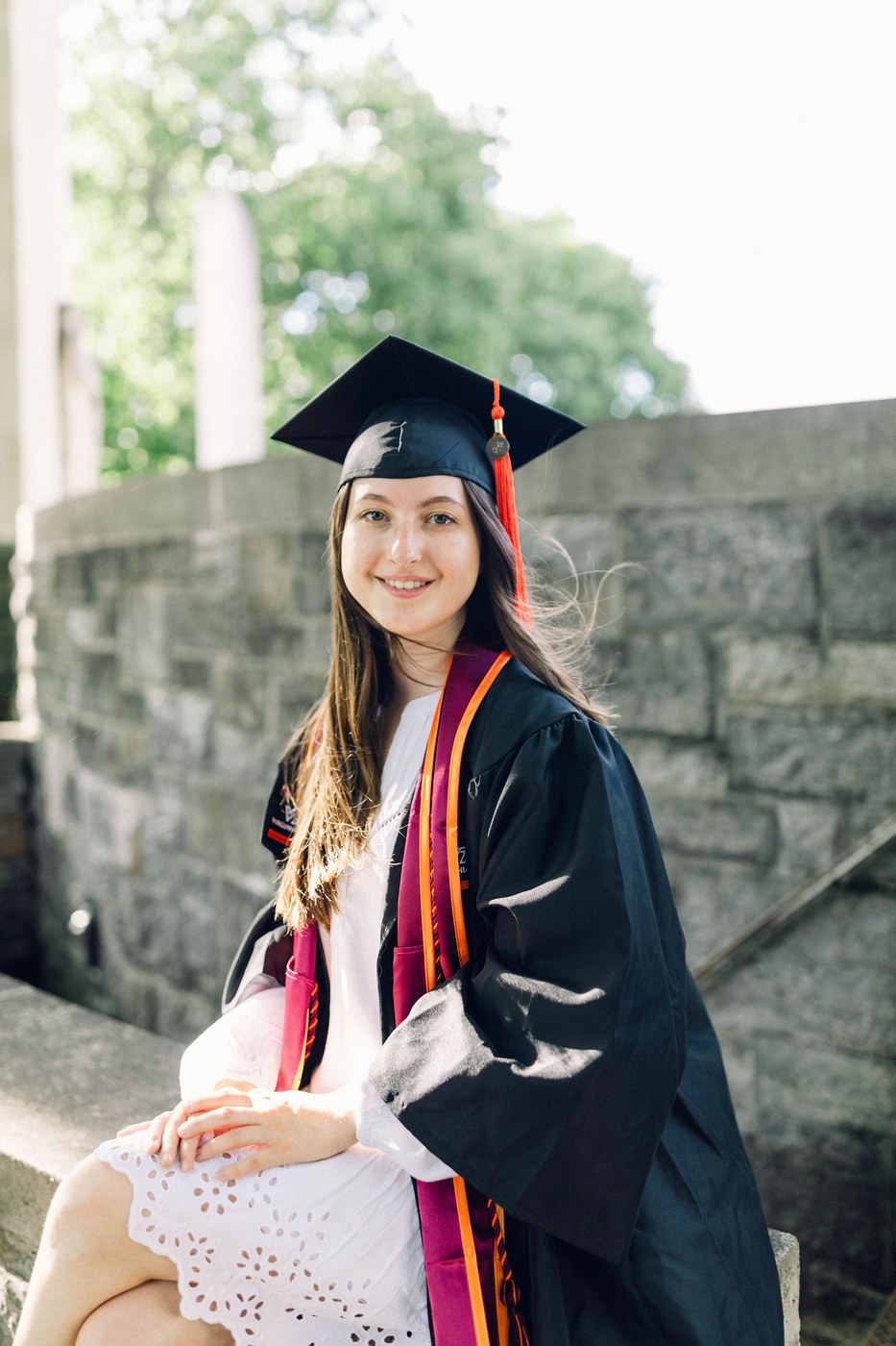 Madeline in her Virginia Tech graduation cap, gown, and stoles, sitting on a stone wall on campus